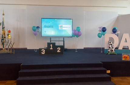 A decorated stage for the Dar Al Fikr Sports Olympics 2025 featuring trophies, a winners’ podium (1–2–3), school-colored balloons, a display screen, and large DAF letters decorated with soccer balls.
